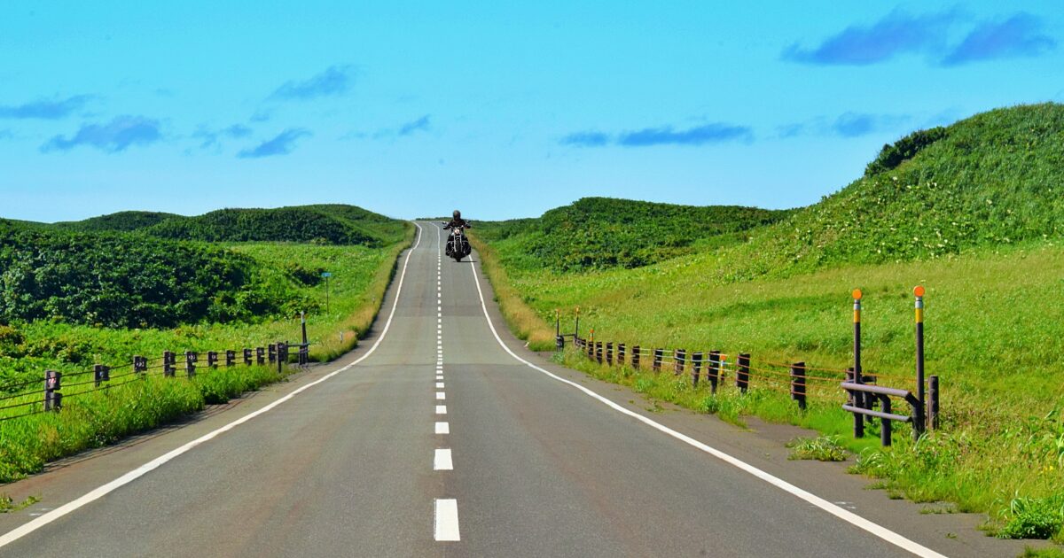 青空と緑の丘に続く一本道を走るツーリングバイク