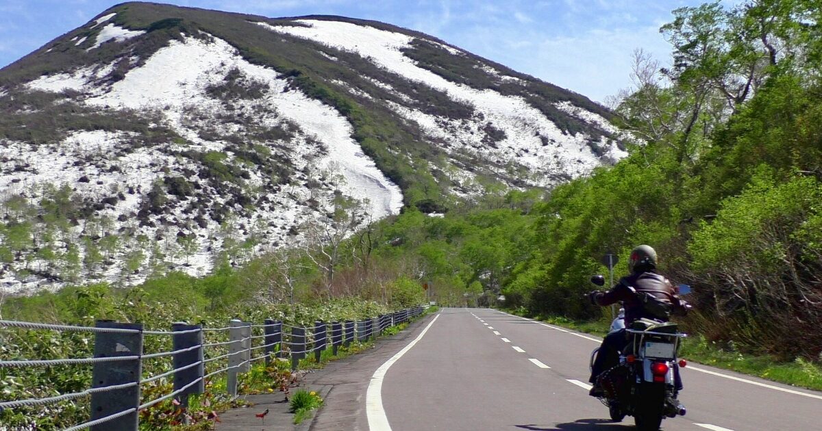 雪山と新緑の中を走るバイクライダーの後ろ姿